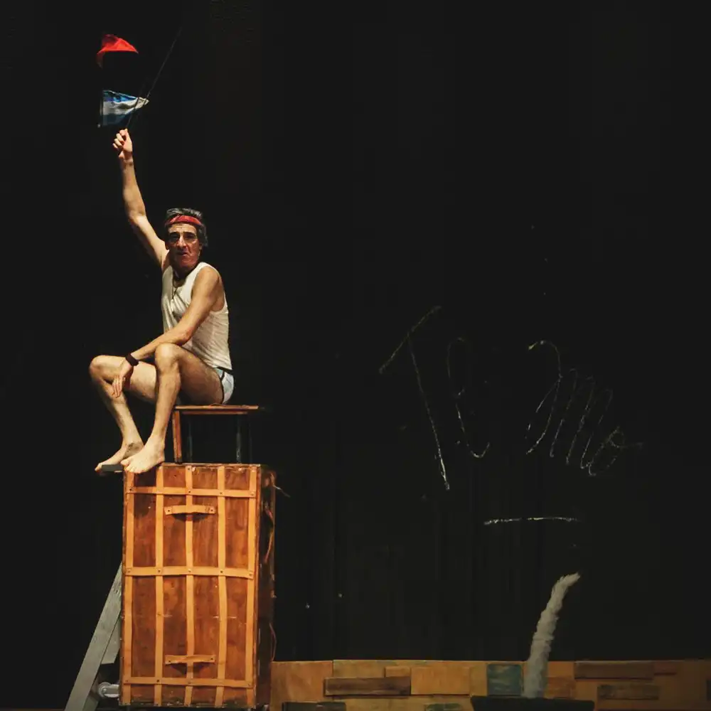 Dayub, en camiseta blanca y calzoncillos, desde lo alto de un armario sostiene una bandera Argentina