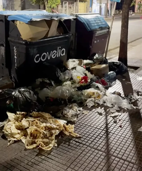 Basura desparramada en una vereda al lado de dos contenedores