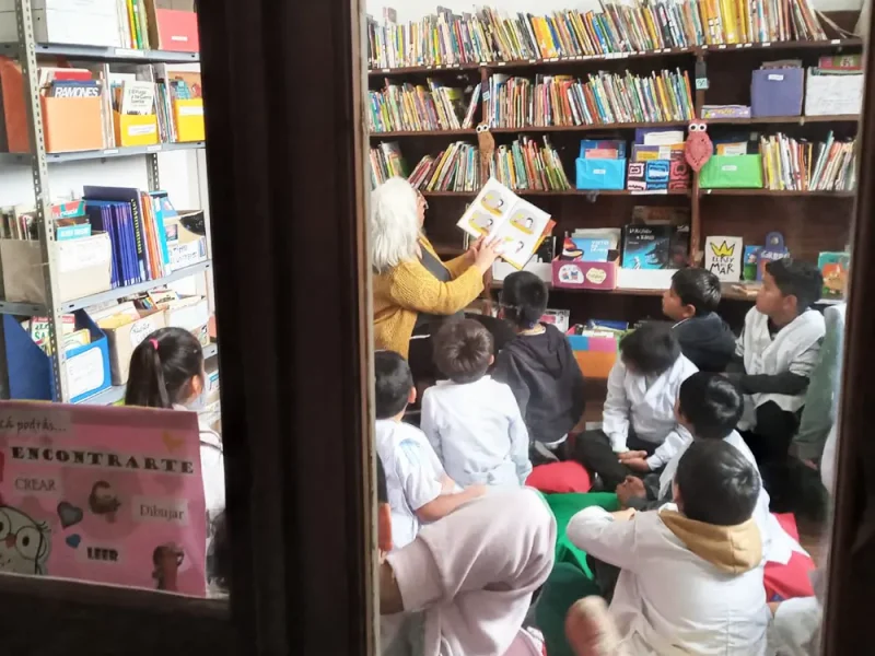 Ni&ntilde;os siguen con la lectura de un libro a su maestra en la blibloteca.